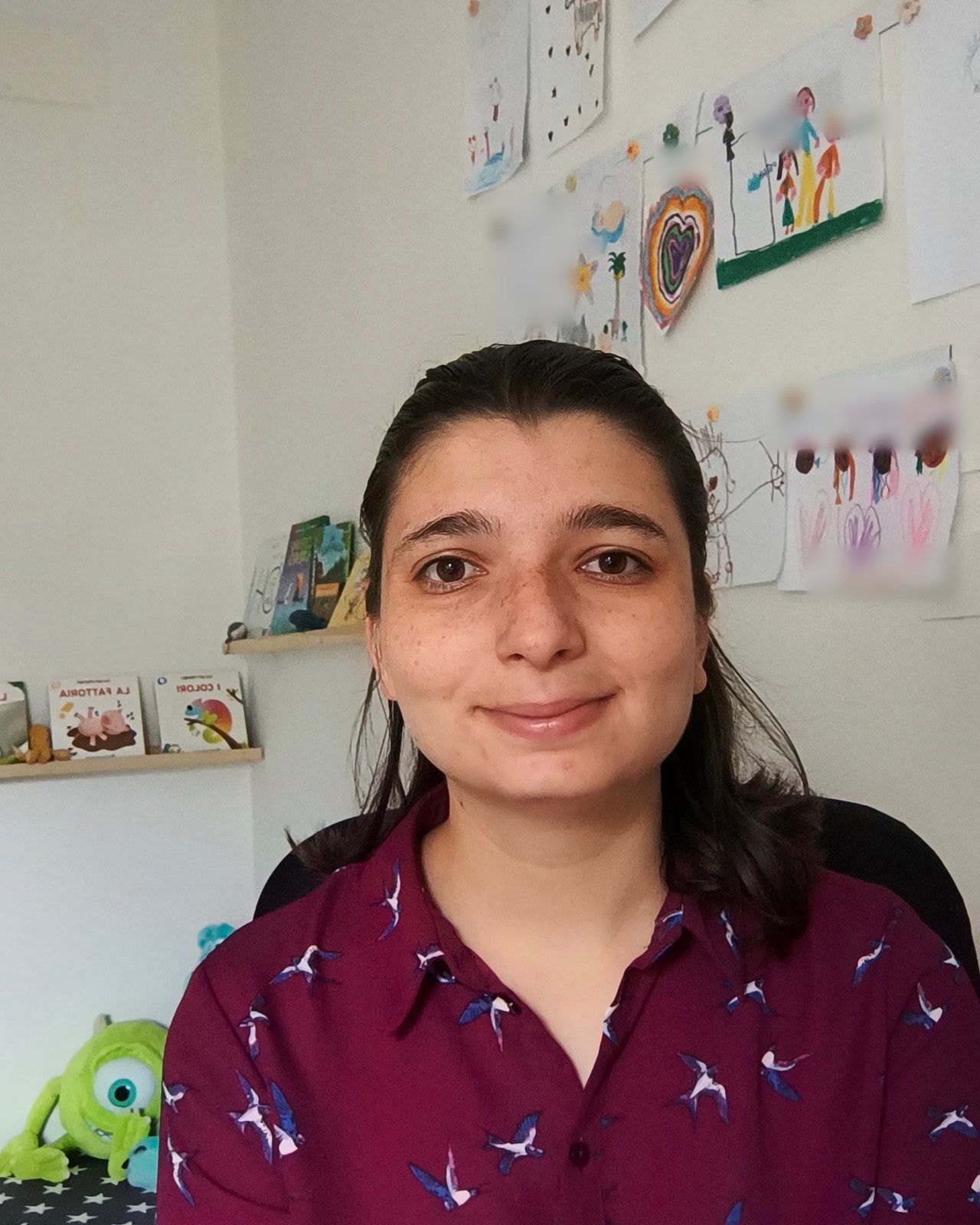 Irene De Simone sorridente nel suo studio, con una camicia bordeaux, davanti ai disegni dei bambini appesi al muro