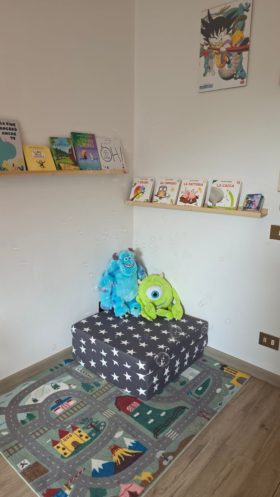 L'angolo lettura: mensole con albi illustrati, due peluche su un pouf a stelle e un tappeto con strade e casette