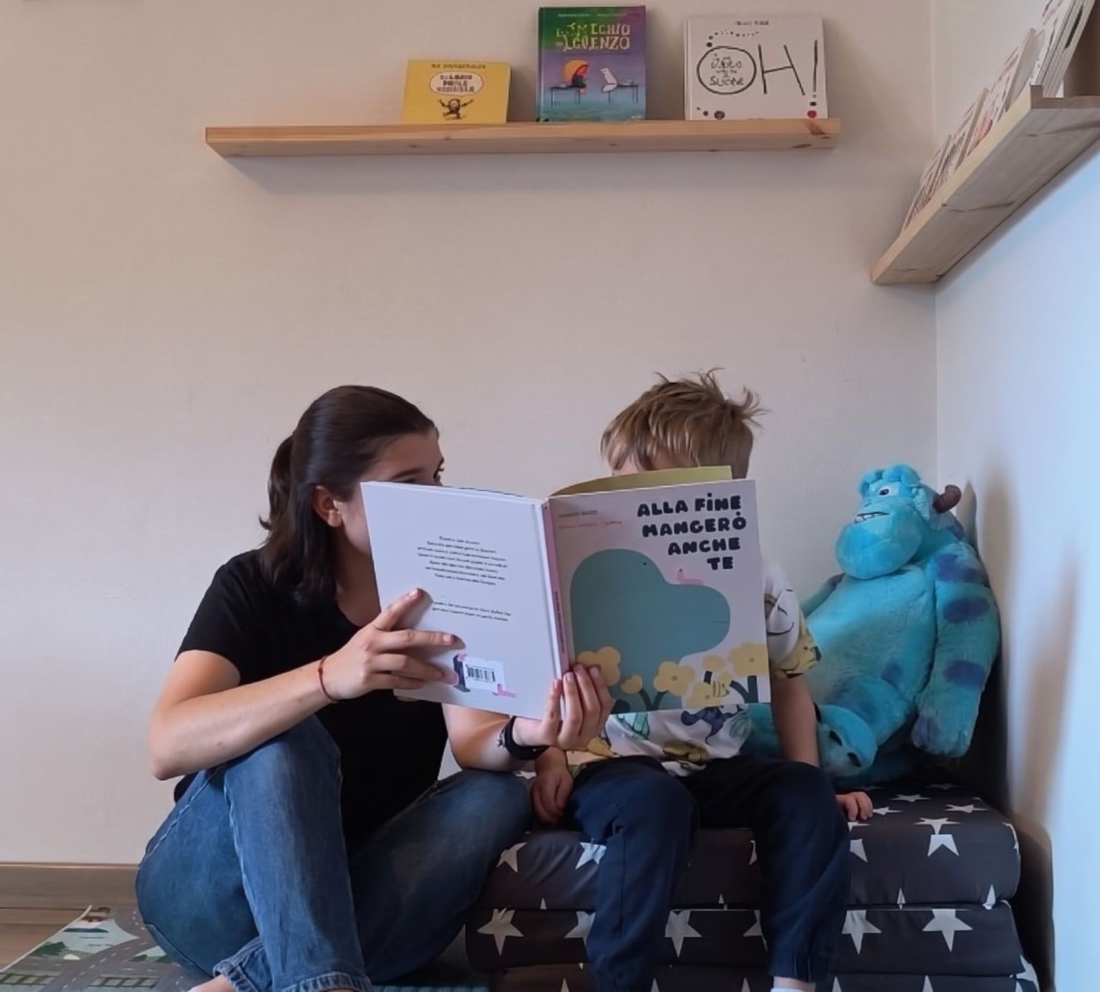 Irene legge un albo illustrato a un bambino nell'angolo lettura dello studio, su un materasso a stelle accanto a un grande peluche