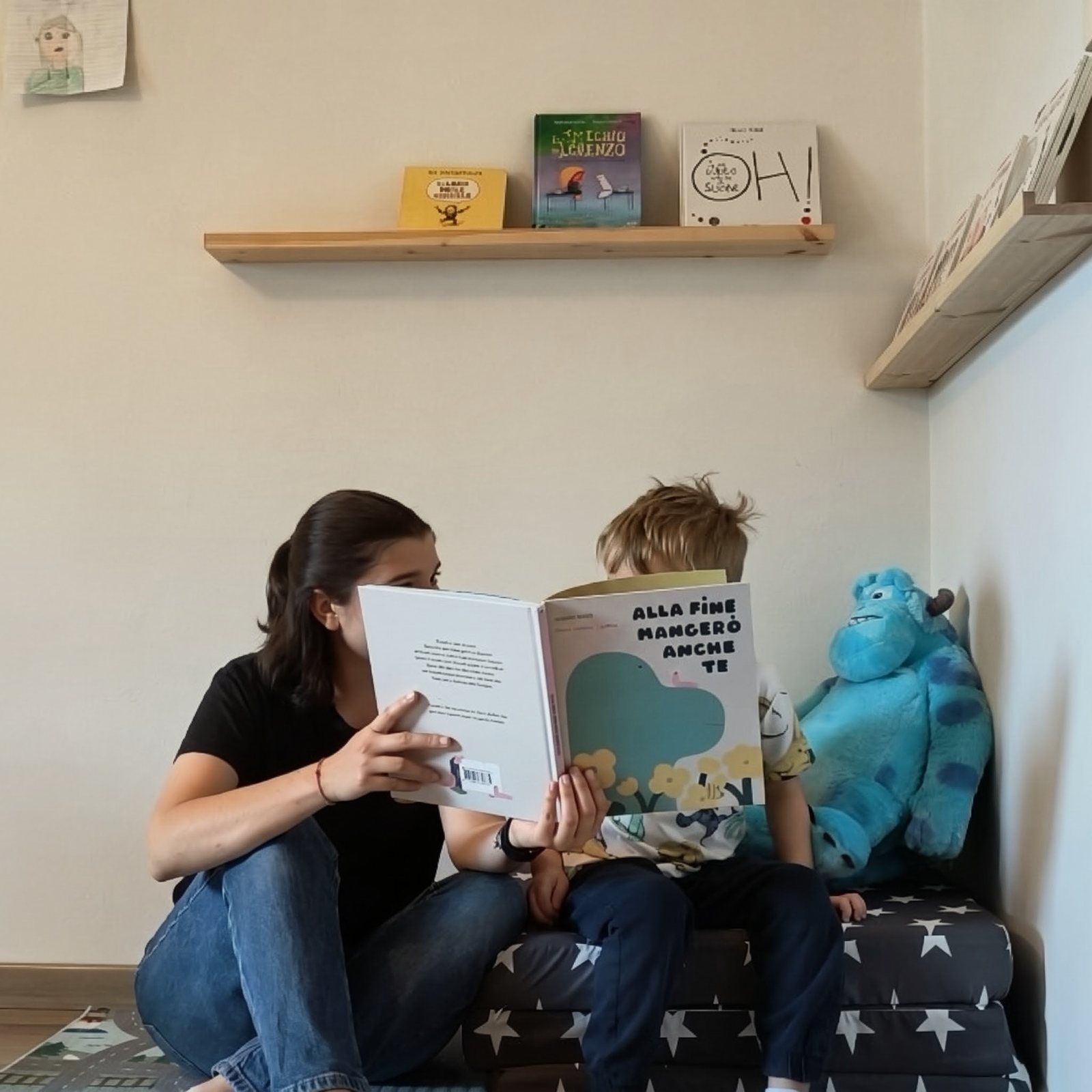 Irene legge un albo illustrato a un bambino nell'angolo lettura dello studio, su un materasso a stelle accanto a un grande peluche