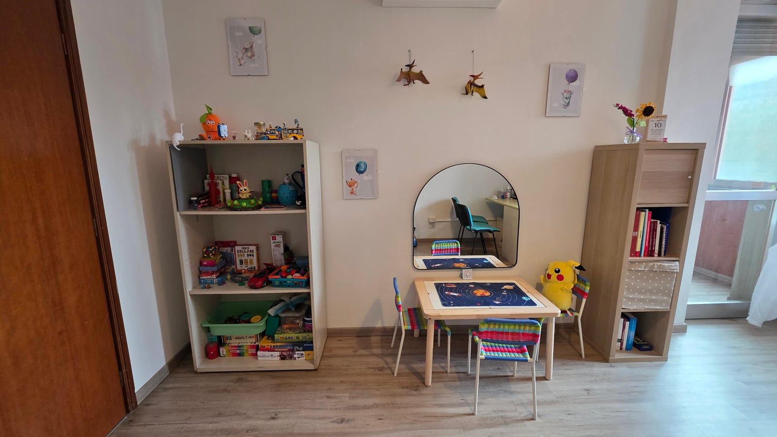 La stanza di terapia: uno scaffale pieno di giochi, il tavolino con le seggioline colorate davanti allo specchio, un peluche di Pikachu e una libreria in legno