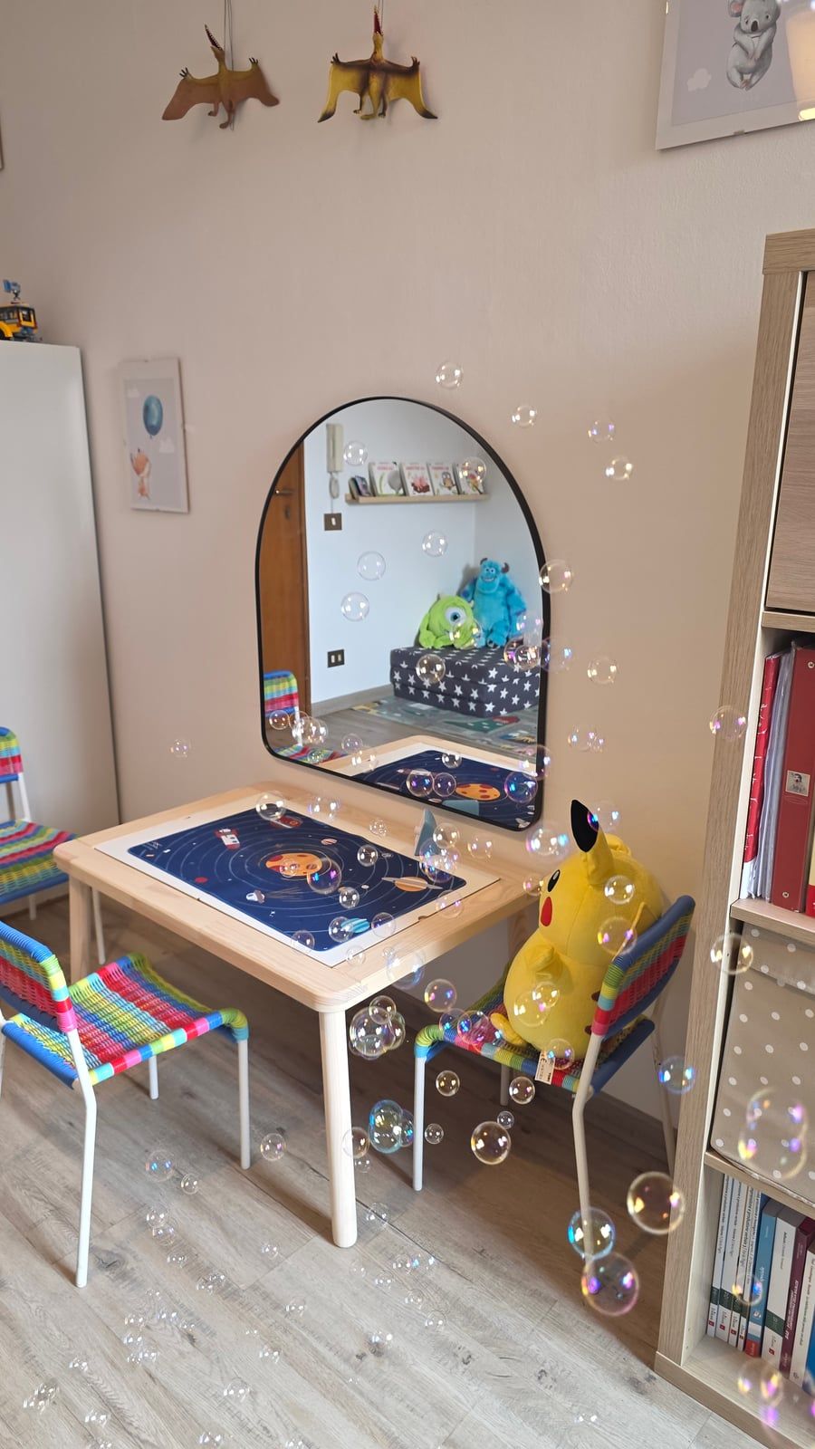 Un tavolino in legno con una tovaglietta dei pianeti e seggioline a righe colorate, uno specchio ad arco alla parete, un peluche di Pikachu su una seggiolina e tante bolle di sapone